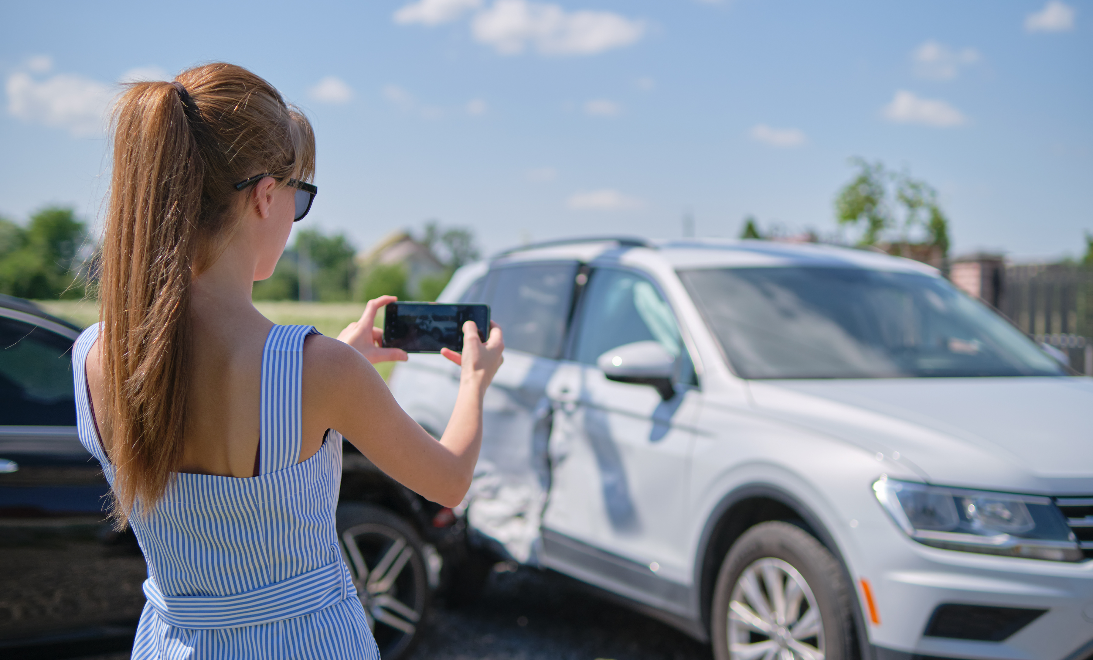 Woman uses phone camera to photograph car accident damage, documenting the vehicle's side impact for an insurance claim.